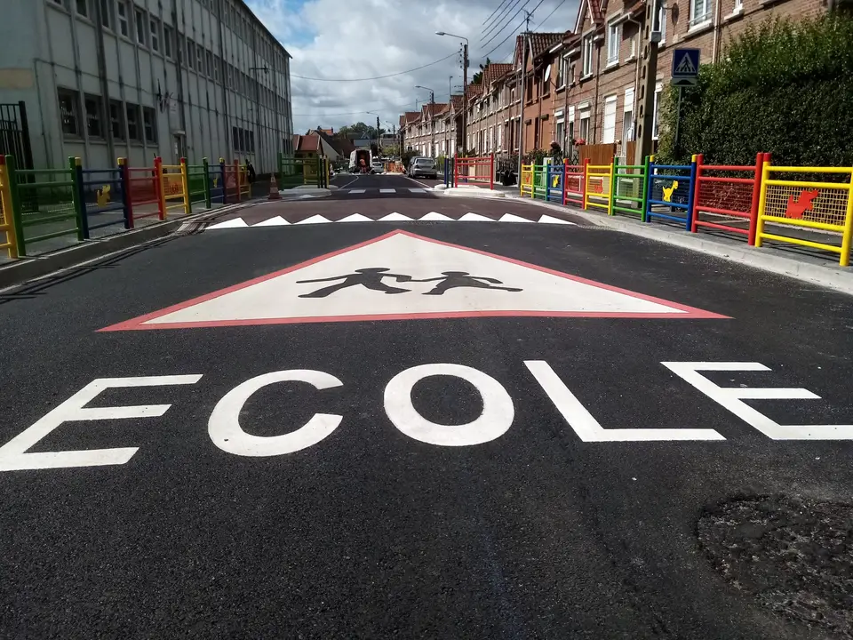 Marquage au sol « ÉCOLE » et triangle à pictogramme d'enfants, rue bordée de barrières colorées
